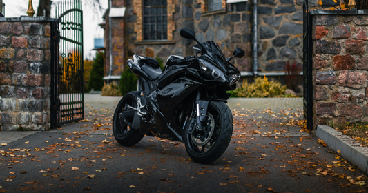 Static black motorcycle parked outside black railing gate, with autumn leaves scattered below on the ground. 
